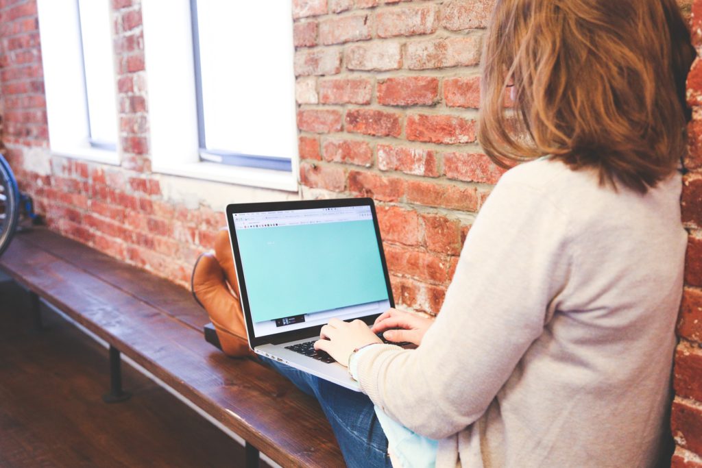The image depicts a woman typing on a laptop on her lap. The woman is wearing blue jeans and a white cardigan, with her back facing the camera.